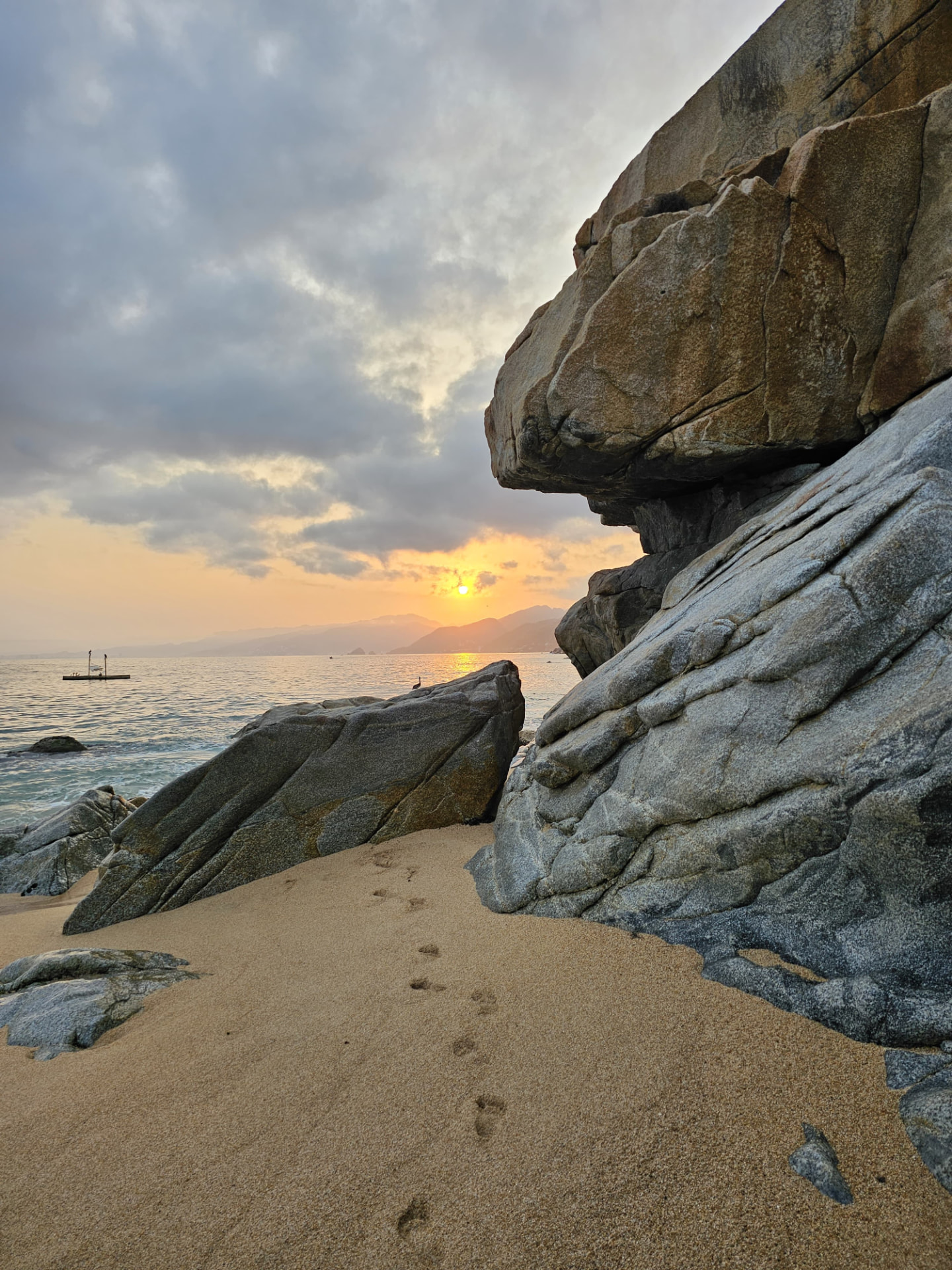 Beach boulders and footprints in the sand as the sun sets over Banderas Bay