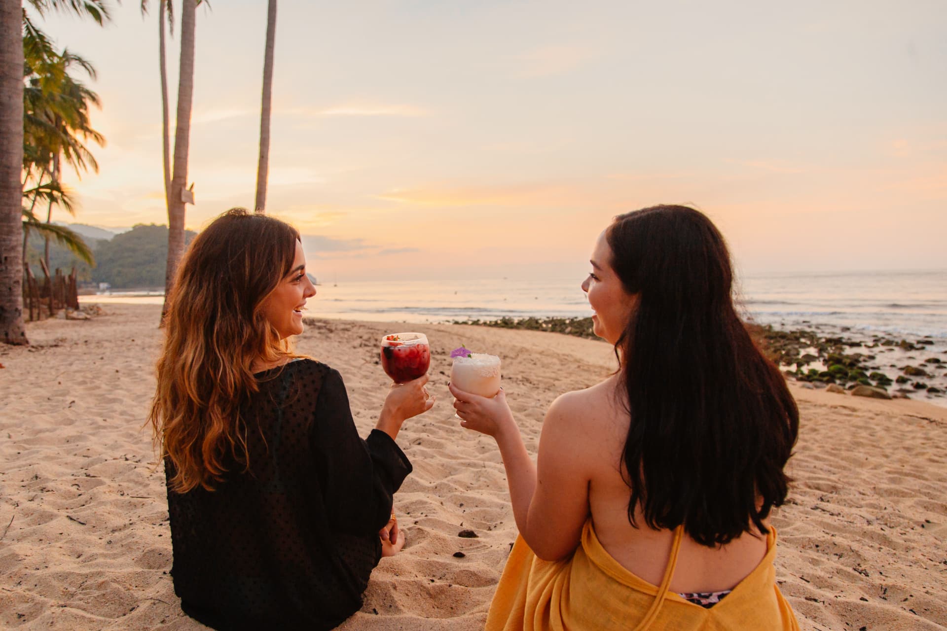 Two friends toasting with cocktails on the beach at sunset